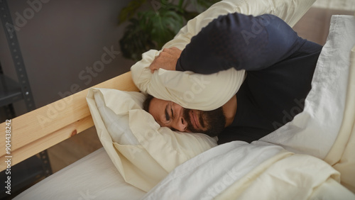 Canvastavla A bearded man expressing frustration in bed, covering his ears with a pillow in a bedroom setting