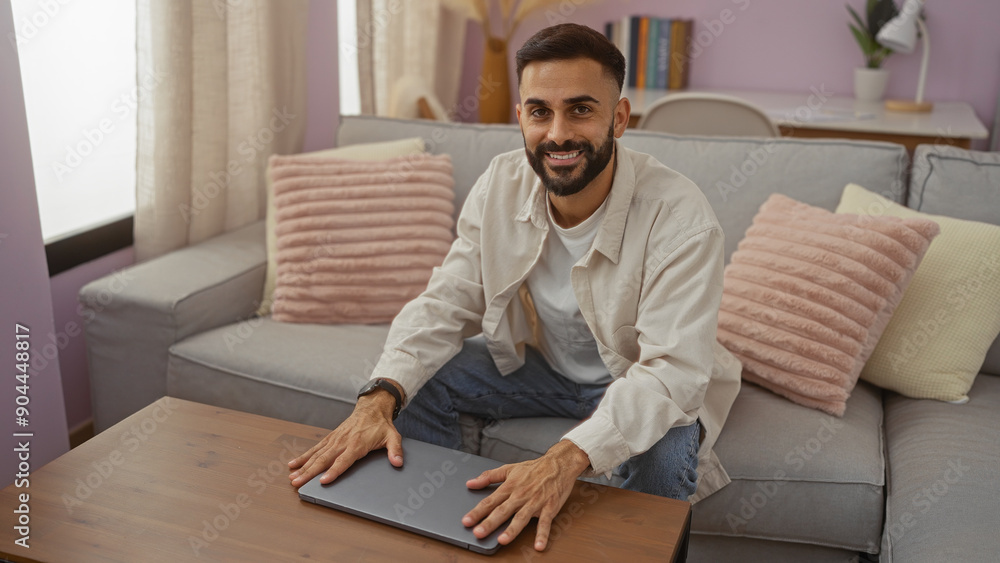 Young, hispanic, man with a beard sitting in a cozy living room with soft pink pillows, smiling at the camera while using his laptop on a wooden table, creating a warm and inviting atmosphere. Young, hispanic, man with a beard sitting in a cozy living room with soft pink pillows, smiling at the camera while using his laptop on a wooden table, creating a warm and inviting atmosphere.