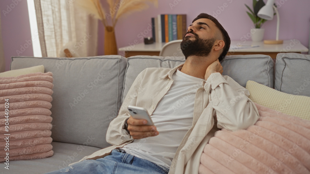 Young man with a beard sitting in a cozy living room, touching his neck in discomfort, while holding a smartphone. Young man with a beard sitting in a cozy living room, touching his neck in discomfort, while holding a smartphone.