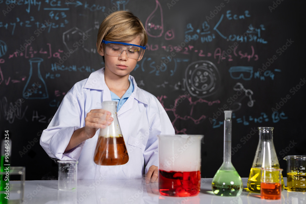 Blonde hair schoolboy in laboratory wear lab coat stand and learn science of chemistry technology in STEM class. The student hold beaker with orange liquid. On the table put many flask. Erudition. Blonde hair schoolboy in laboratory wear lab coat stand and learn science of chemistry technology in STEM class. The student hold beaker with orange liquid. On the table put many flask. Erudition.
