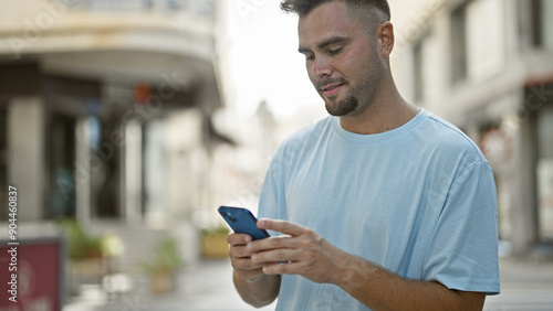 A young hispanic man casually using a smartphone on a sunny urban street, exemplifying modern connectivity and city life.