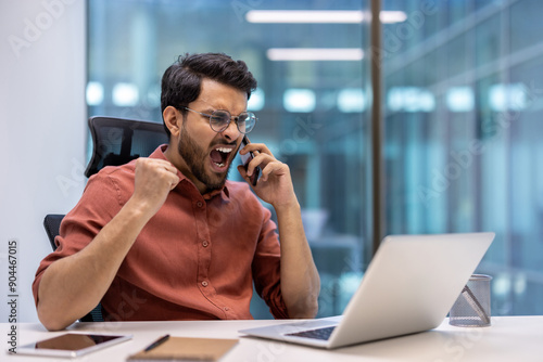 Angry businessman shouting on phone at office desk expressing frustration. Young professional arguing call with clenched fist, laptop, and tablet on desk. Office environment with blurred background.