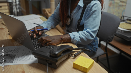 An african american woman working as a detective types on a laptop in a police station office.