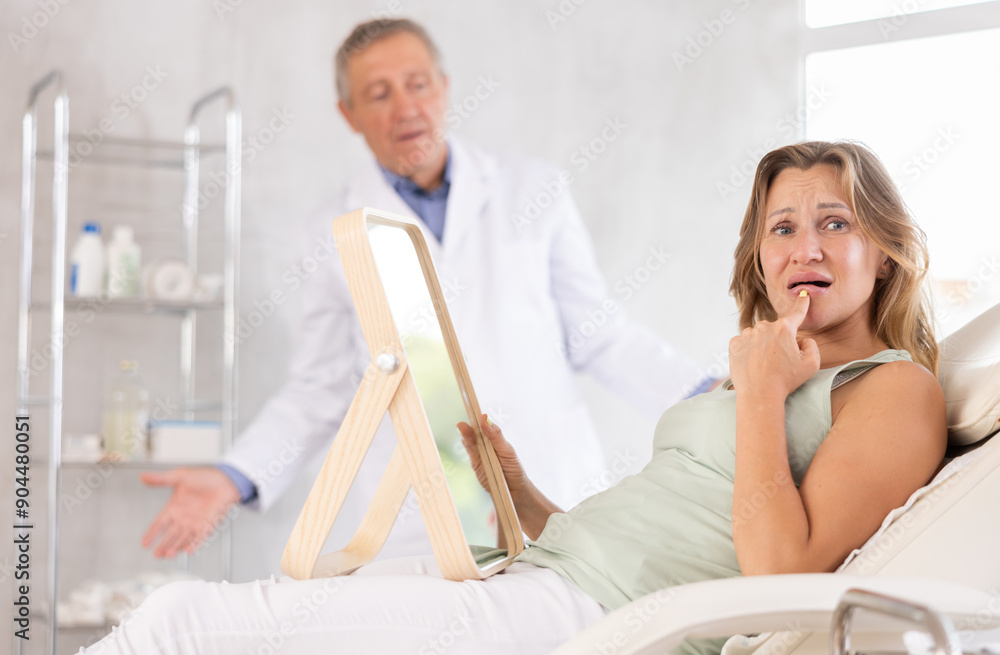 Obraz premium Upset female patient behaving discontentedly lying on medical chair with mirror in hands in aesthetic clinic