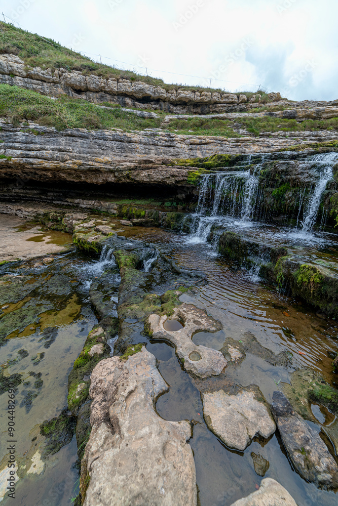 Obraz premium Picturesque waterfall flowing over layered rock formations with lush green moss in Cantabria, Spain. Ideal for nature, travel, and landscape themes.