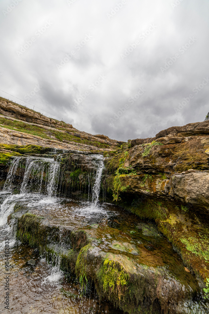 Obraz premium Picturesque waterfall flowing over layered rock formations with lush green moss in Cantabria, Spain. Ideal for nature, travel, and landscape themes.