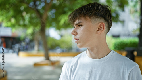 Caucasian teenage boy with blond hair in profile, wearing a white shirt, contemplating in a green, outdoor urban park.
