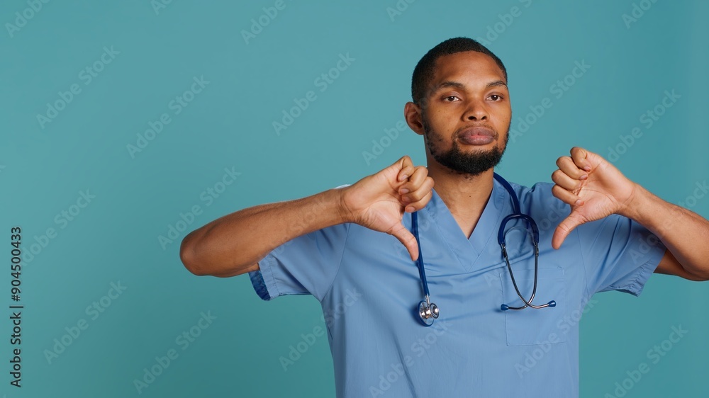 Sad african american male nurse showing thumbs down signs while at work, isolated over studio backdrop. Unhappy healthcare employee wearing scrubs doing disapproval gestures, camera A Sad african american male nurse showing thumbs down signs while at work, isolated over studio backdrop. Unhappy healthcare employee wearing scrubs doing disapproval gestures, camera A