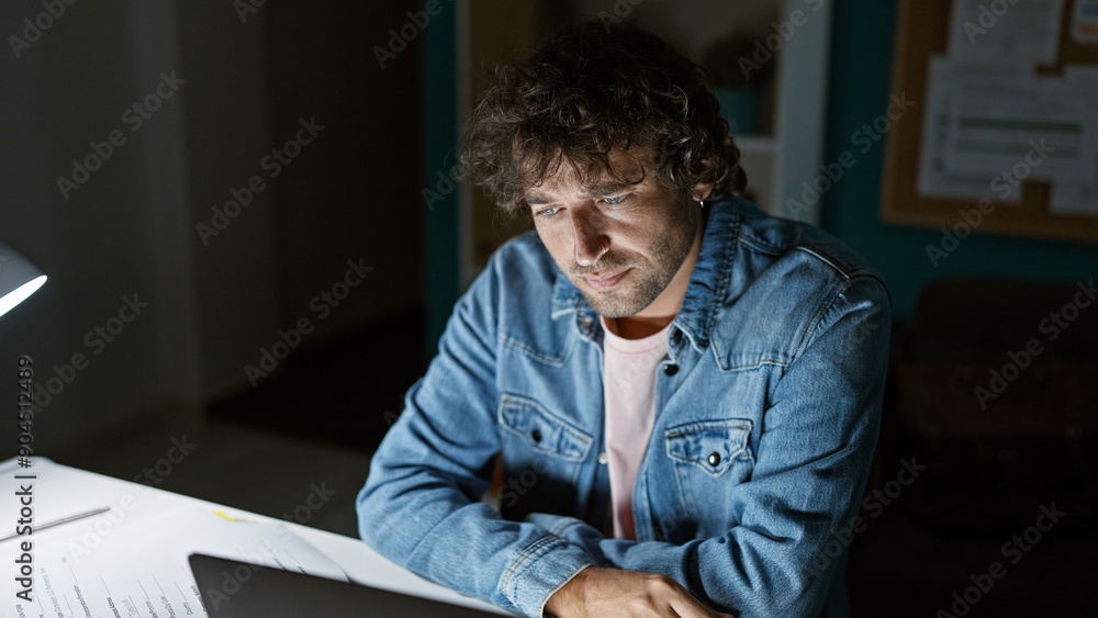 A focused hispanic man working late at his office desk, illuminated by a desk lamp in a dark room. A focused hispanic man working late at his office desk, illuminated by a desk lamp in a dark room.