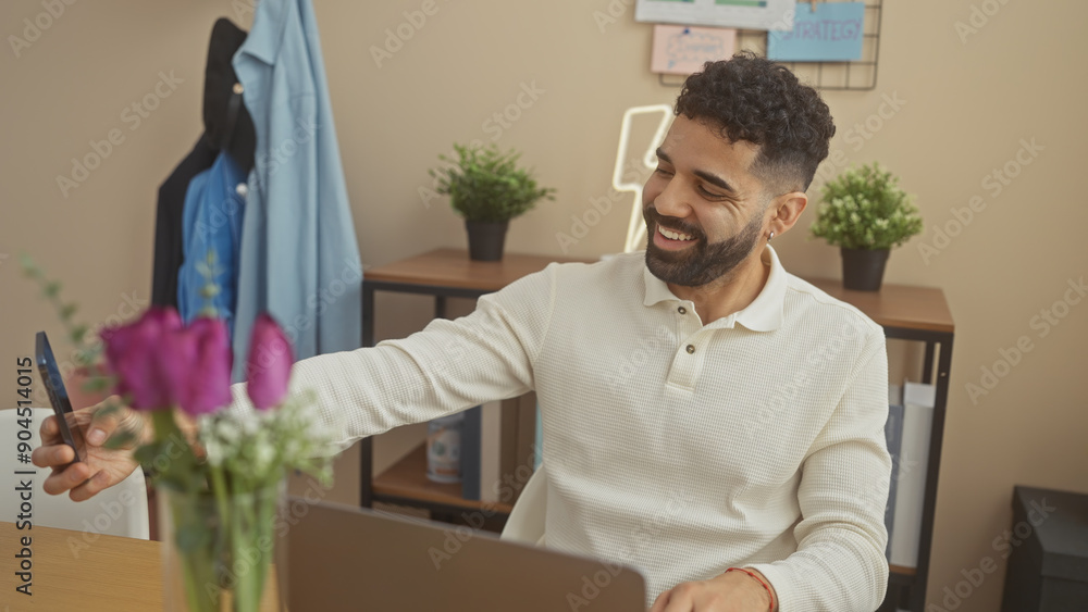 Fototapeta premium A cheerful young hispanic man takes a selfie in a modern home office with decor and laptop.