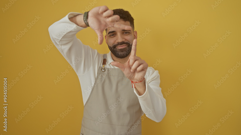 A young hispanic man in a beige apron framing his face with his hands against a yellow background. A young hispanic man in a beige apron framing his face with his hands against a yellow background.