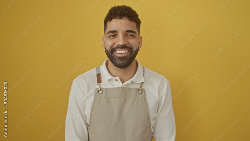 Handsome hispanic man with beard wearing apron smiling against yellow wall Handsome hispanic man with beard wearing apron smiling against yellow wall