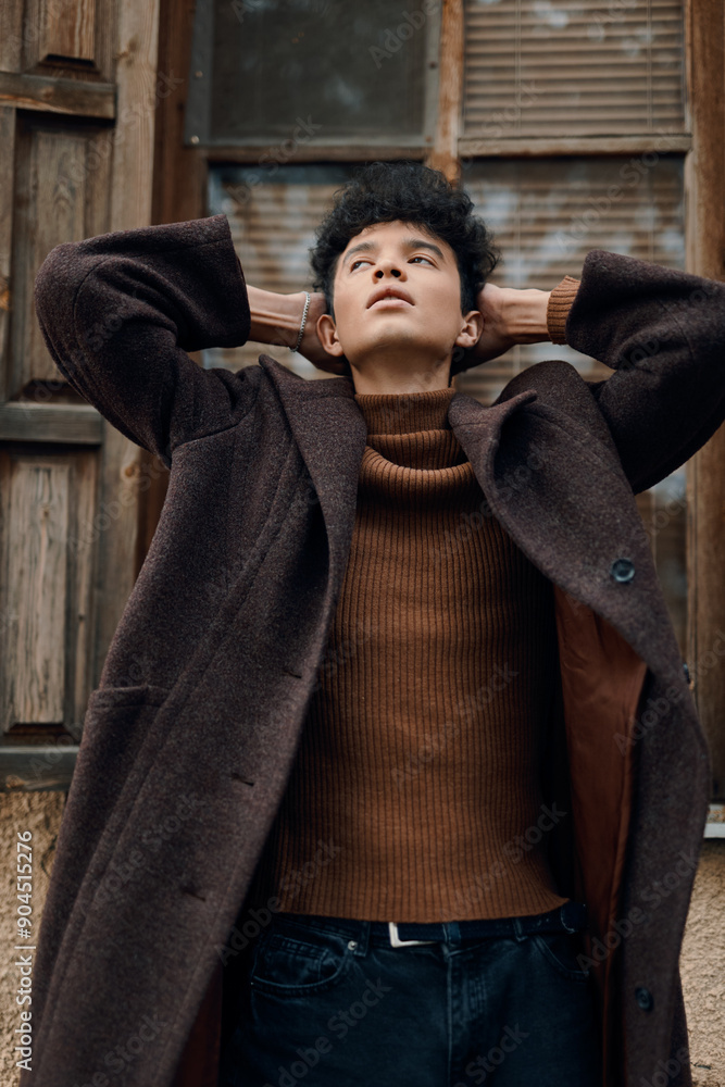Stylish young man in brown coat and black turtleneck leaning against window with hands on head, looking at camera Stylish young man in brown coat and black turtleneck leaning against window with hands on head, looking at camera