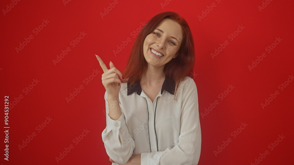 Young, attractive redhead woman smiling and pointing upwards, isolated against a red background. Young, attractive redhead woman smiling and pointing upwards, isolated against a red background.