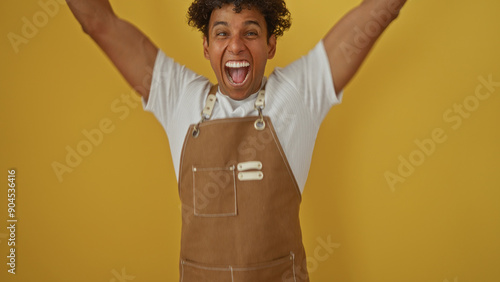 Young man in a brown apron exuberantly celebrating over an isolated yellow background. photo