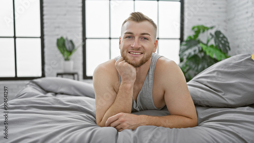 A young bearded man smiling in a bright bedroom with white walls and a plant