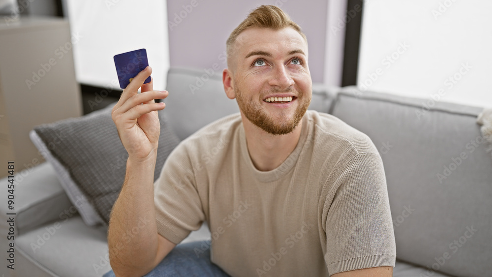 Attractive young man holding credit card indoors, looking optimistic in a casual apartment setting. Attractive young man holding credit card indoors, looking optimistic in a casual apartment setting.