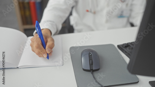 A man in a white coat writing notes with a computer in a modern clinic office, depicting a healthcare professional at work. photo