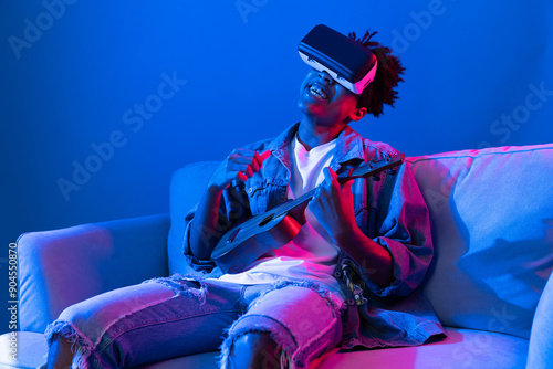 Foto Young African American singer using through VR glasses enjoy singing song with guitar playing on meta blue neon light wall virtual reality metaverse fantasy technology sitting on sofa