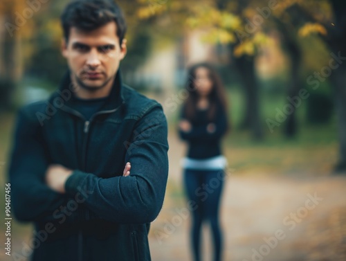 Cena pós-briga de casal: homem de braços cruzados em primeiro plano, mulher ao fundo com expressão angustiada. Reflete distanciamento emocional e tensão após discussão photo