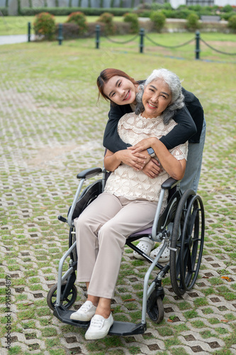 A lovely granddaughter is hugging her grandma, who is in a wheelchair, while walking her in a park.