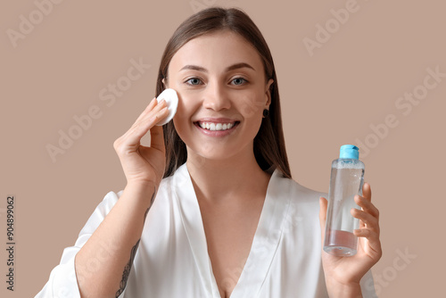 Beautiful young woman applying toner with cotton pad on beige background, closeup photo