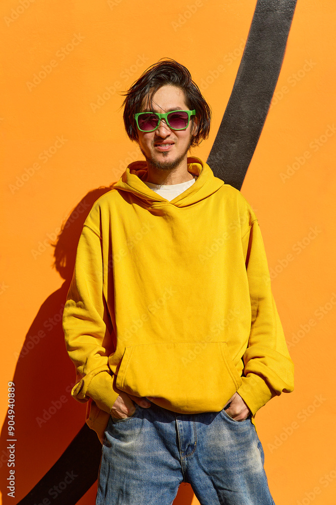 An Asian guy in stylish green glasses stands against a yellow wa An Asian guy in stylish green glasses stands against a yellow wa