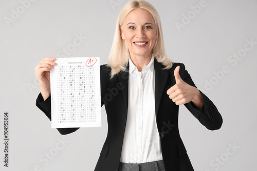 Mature female teacher with results of exam showing thumb-up on light background