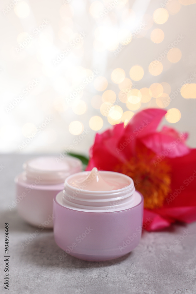 Jars of creams and peony flower on gray table against light background with blurred lights Jars of creams and peony flower on gray table against light background with blurred lights