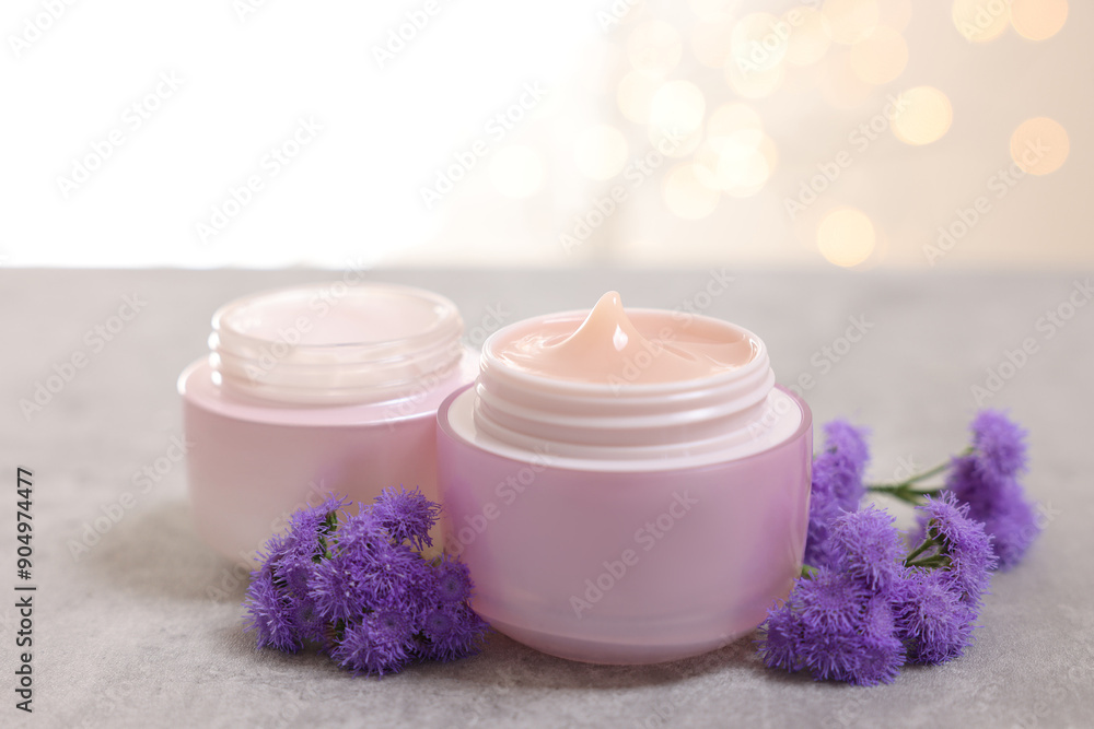 Jars of creams and ageratum flowers on gray table against light background with blurred lights, closeup Jars of creams and ageratum flowers on gray table against light background with blurred lights, closeup