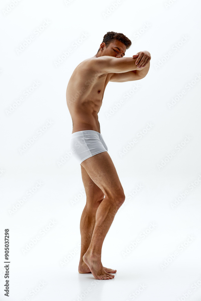 Obraz premium Young man wearing white boxer briefs stretches his arm, showcasing his sculpted body and serious expression against white studio background. Concept of body care, male health, natural beauty, youth