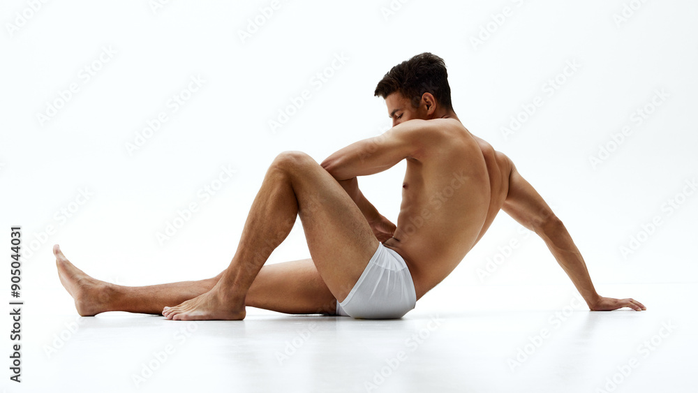 Obraz premium Fit young man in white boxer briefs sits on floor, leaning back on one arm with thoughtful expression against white studio background. Concept of body care, male health, natural beauty, youth. Ad