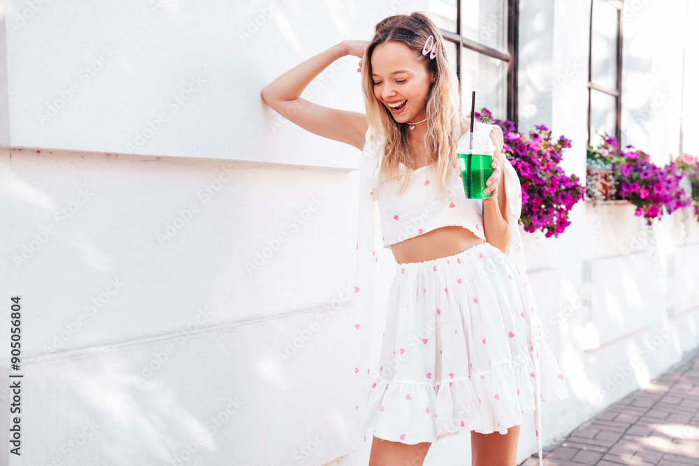 Young beautiful smiling hipster woman in trendy summer clothes. Sexy carefree model posing in the street. Holding and drinking fresh vegetable cocktail drink in plastic cup with straw. In sunny day Young beautiful smiling hipster woman in trendy summer clothes. Sexy carefree model posing in the street. Holding and drinking fresh vegetable cocktail drink in plastic cup with straw. In sunny day
