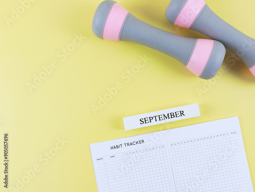  flat lay of habit tracker book, wooden calendar September, gray pink dumbbells,  on yellow  background with copy space. Self development and active lifestyle concept.