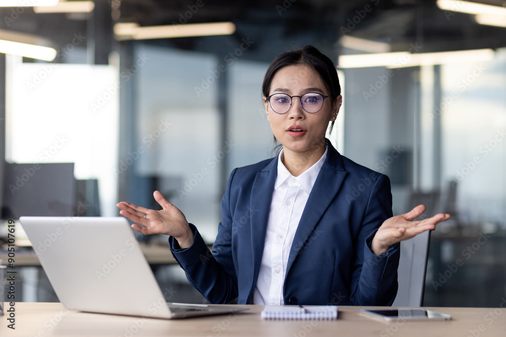 Confused businesswoman in formal attire working on laptop while looking at camera in modern office. Woman appears perplexed, raising hands, with notebook and smartphone on desk. Confused businesswoman in formal attire working on laptop while looking at camera in modern office. Woman appears perplexed, raising hands, with notebook and smartphone on desk.