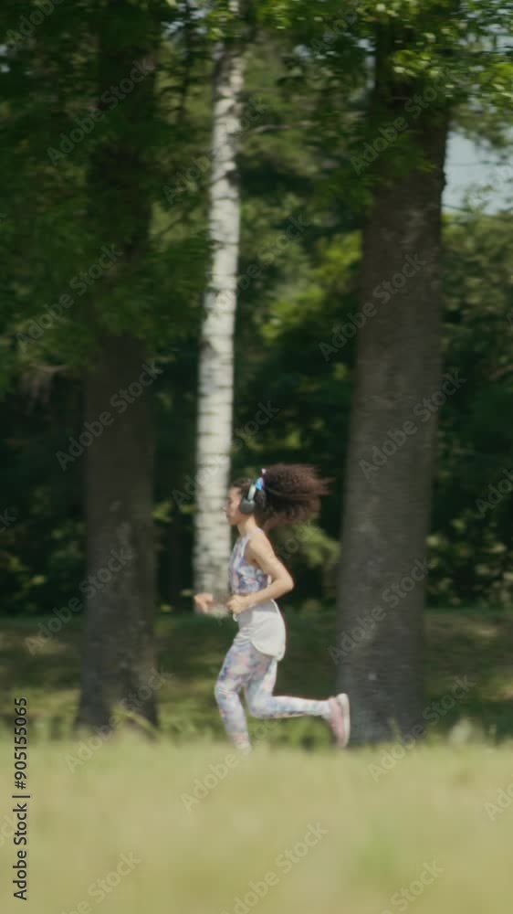 Vertical shot of young woman in headphones and sportswear running in park with lush trees and green grass while training outdoors on summer day Vertical shot of young woman in headphones and sportswear running in park with lush trees and green grass while training outdoors on summer day