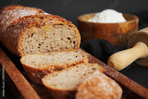 Fresh sliced ​​bread on a wooden tray next to a bowl of flour and a wooden rolling pin on a dark background