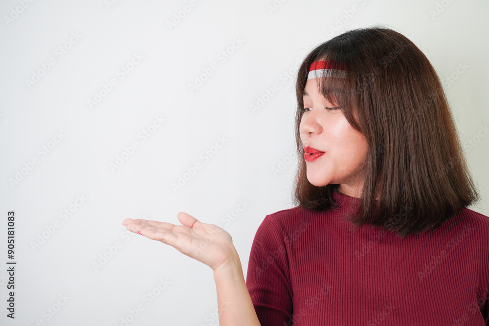 Surprised young asian woman wearing Indonesian flag headband, amazed expression with hands open gesture carrying something, isolated over white background. Concept for Indonesian Independence Day.