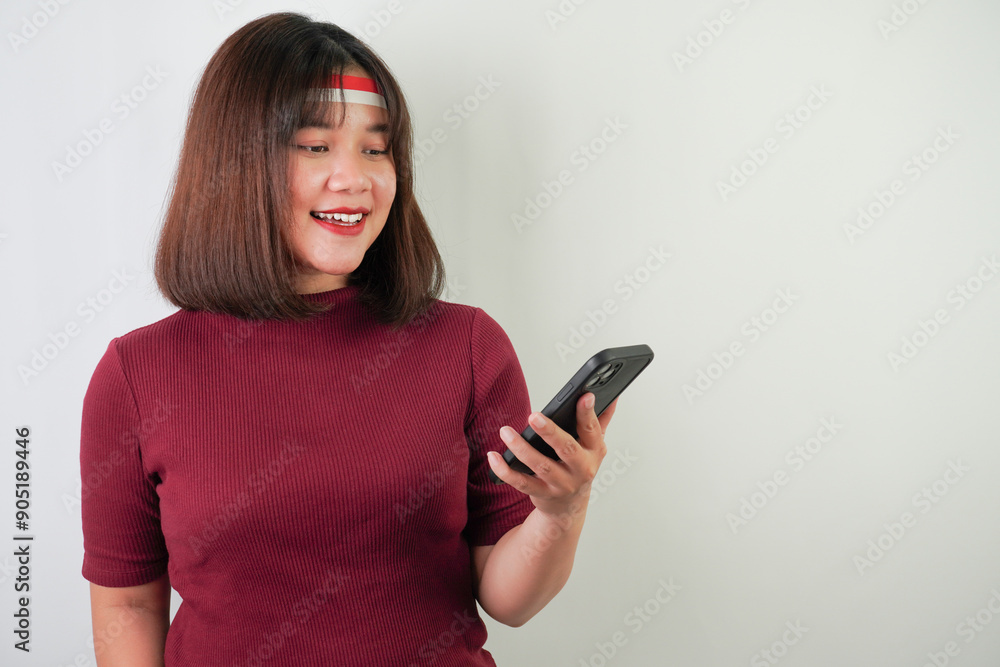 Excited young asian woman wearing Indonesian Flag headband is watching vertical smartphone screen for advertising, clenched fist gesture, isolated over white background. Indonesian Independence Day. Excited young asian woman wearing Indonesian Flag headband is watching vertical smartphone screen for advertising, clenched fist gesture, isolated over white background. Indonesian Independence Day.