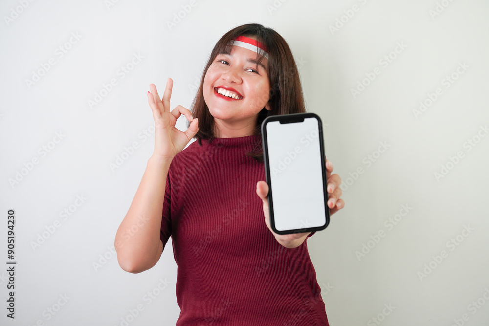 Naklejka premium Smiling young asian woman wearing Indonesian Flag headband is showing vertical blank smartphone screen for advertising with okay gesture, isolated over white background. Indonesian Independence Day.