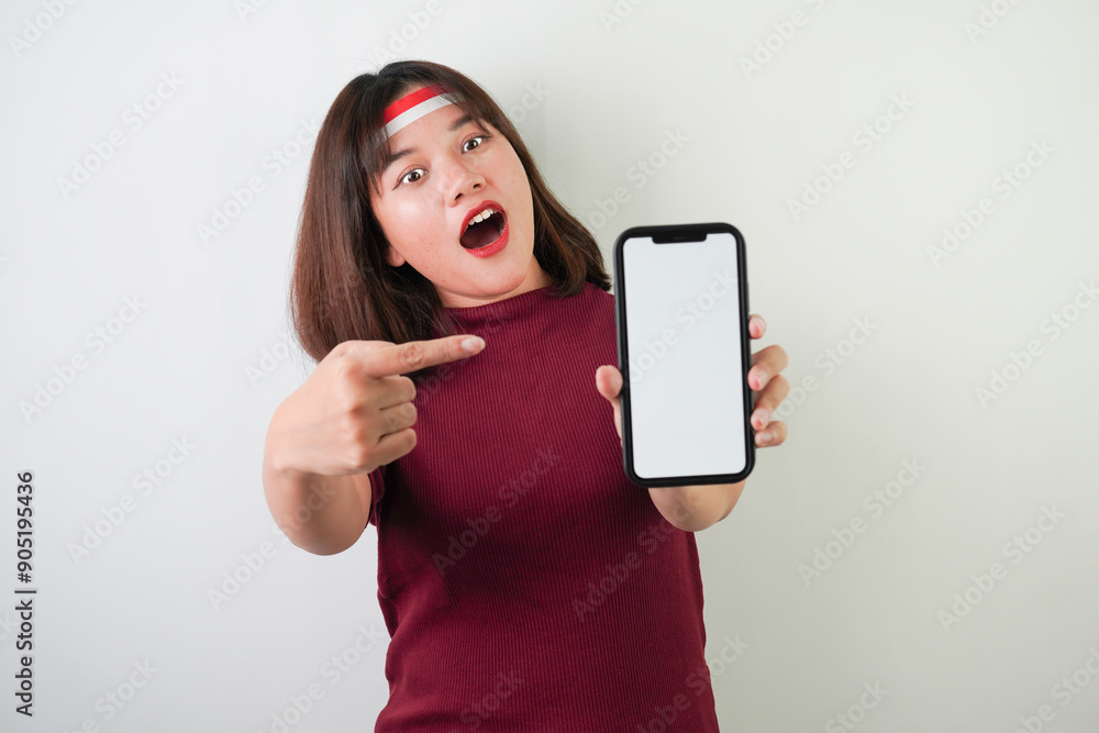 Fototapeta premium Surprised amazed young asian woman wearing Indonesian Flag headband is pointing vertical blank smartphone screen for advertising, isolated over white background. Indonesian Independence Day.
