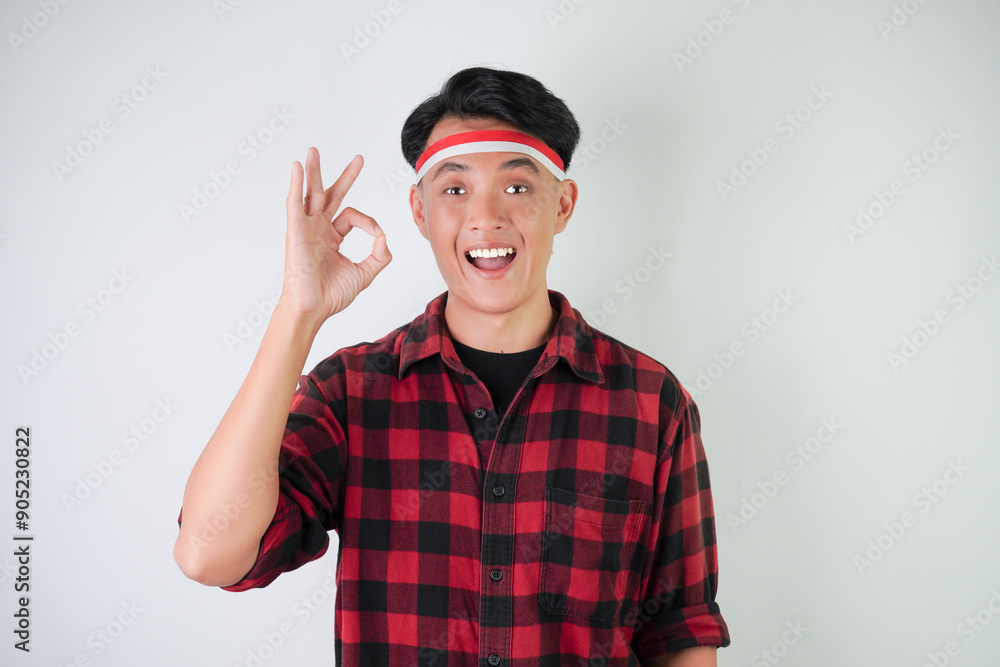 Excited young asian man wearing Indonesian flag headband, smiling expression, hand raised with okay gesture, isolated over white background. Concept for Indonesian Independence Day. Excited young asian man wearing Indonesian flag headband, smiling expression, hand raised with okay gesture, isolated over white background. Concept for Indonesian Independence Day.