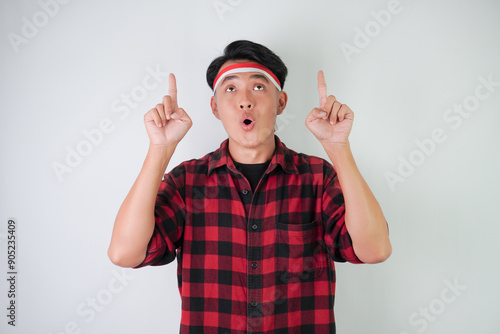 Surprised amazed young asian man wearing Indonesian flag headband and pointing fingers on copy space upside, isolated over white background. Concept for Indonesian Independence Day.