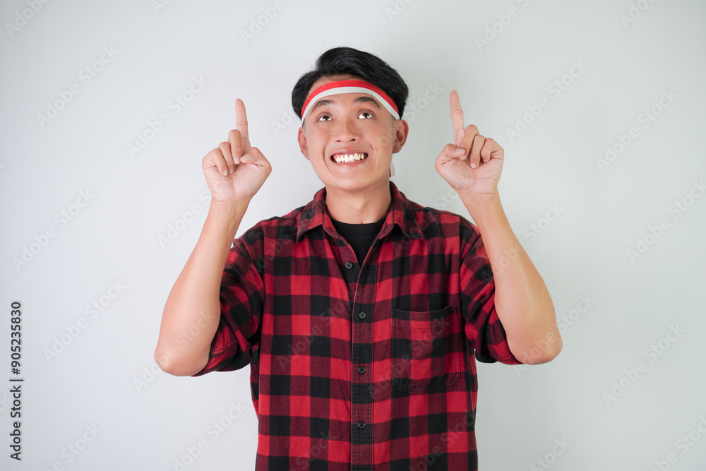 Surprised amazed young asian man wearing Indonesian flag headband and pointing fingers on copy space upside, isolated over white background. Concept for Indonesian Independence Day. Surprised amazed young asian man wearing Indonesian flag headband and pointing fingers on copy space upside, isolated over white background. Concept for Indonesian Independence Day.