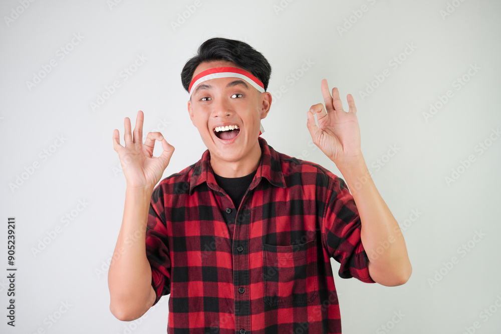 Excited young asian man wearing Indonesian flag headband, smiling expression, hand raised with okay gesture, isolated over white background. Concept for Indonesian Independence Day. Excited young asian man wearing Indonesian flag headband, smiling expression, hand raised with okay gesture, isolated over white background. Concept for Indonesian Independence Day.