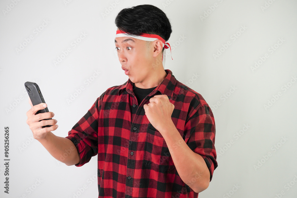 Surprised shocked young asian man wearing Indonesian Flag headband is watching vertical smartphone screen for advertising, isolated over white background. Indonesian Independence Day. Surprised shocked young asian man wearing Indonesian Flag headband is watching vertical smartphone screen for advertising, isolated over white background. Indonesian Independence Day.