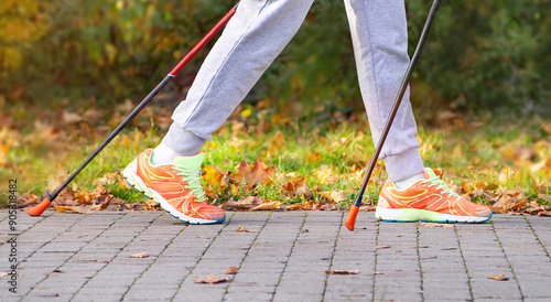 Legs of woman in sport wear and walking sticks for popular nordic walking in the autumnal park. Scandinavian trekking sticks hilking, exercising, healthy life style. Sneakers shoes. Copy space. photo