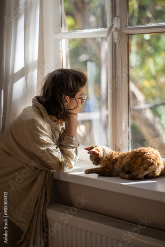 Lonely upset young woman leaning on windowsill near cat need psychologies help. Outcast by friends teen girl feeling frustration sadly looking at window loving cat trying emotional support to owner. photo