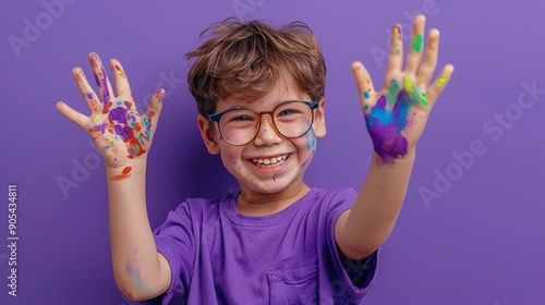 A young boy with glasses, dressed in a purple shirt, is holding his hands up, covered in paint with a proud and happy smile. The bright purple background enhances the colorful and artistic mood