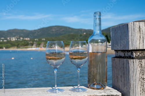 Bottle and glass of cold rose wine from Provence and wooden yacht boota pier on white sandy beach Plage de Pampelonne near Saint-Tropez, summer vacation in France photo